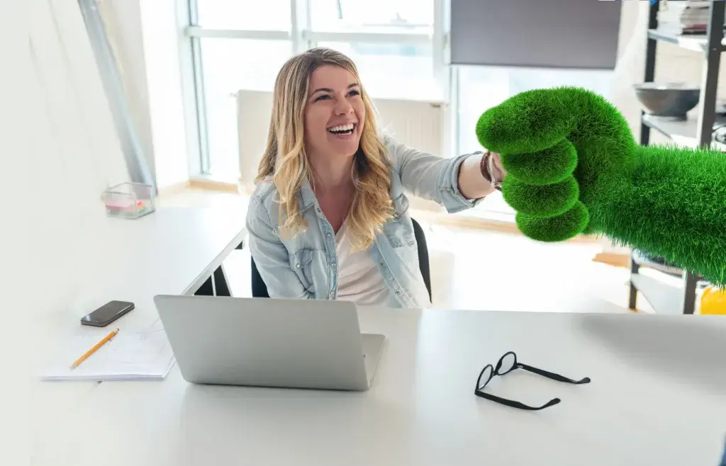 A woman sitting at a desk smiles and extends her hand for a shake, greeted by a large, green, oversized hand, creating a playful and lively atmosphere in an office setting.