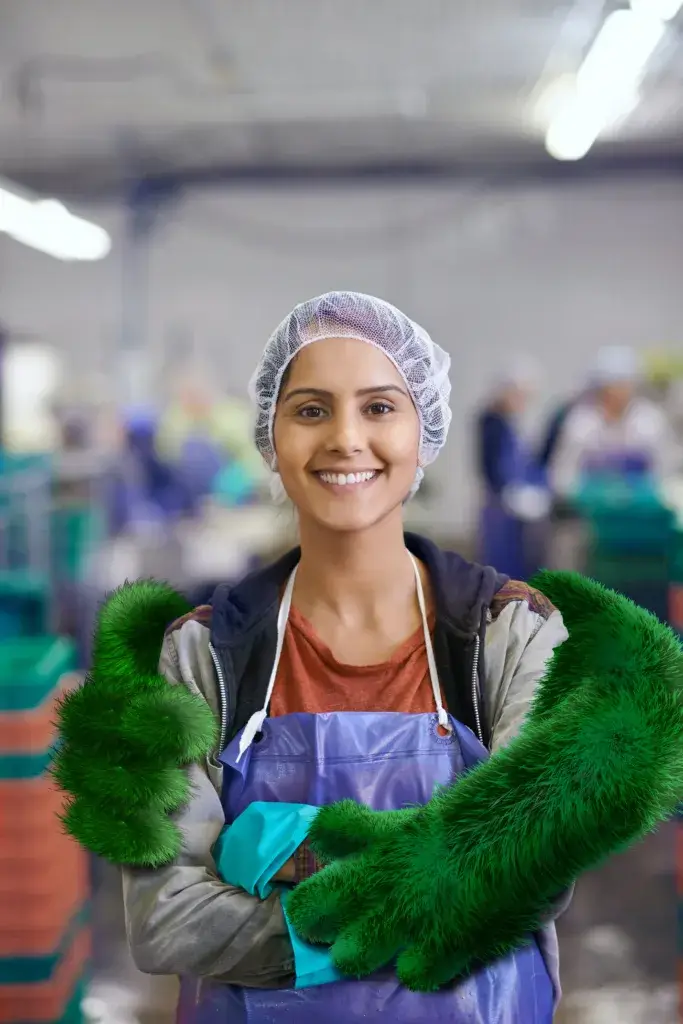 A smiling woman wearing a hairnet and apron poses with her arms crossed, showcasing green gloves. The background shows a busy workspace, indicating an active environment.