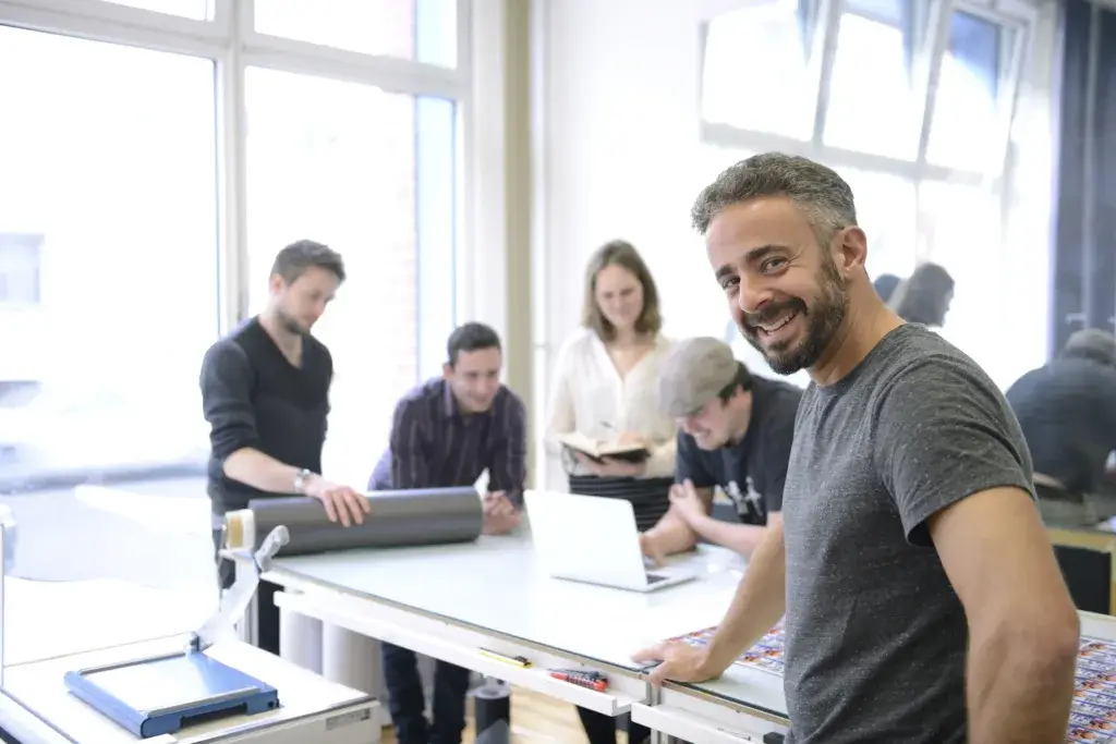 A diverse group of five people collaborates in a bright workspace. One man smiles at the camera while others engage with a laptop and materials on a table, suggesting teamwork and creativity in a professional setting.